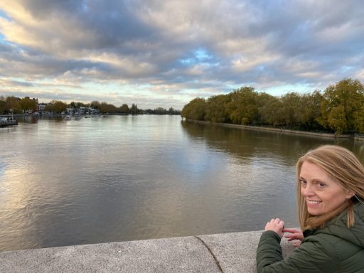 A woman with long hair smiles by a river, surrounded by trees and cloudy skies.
